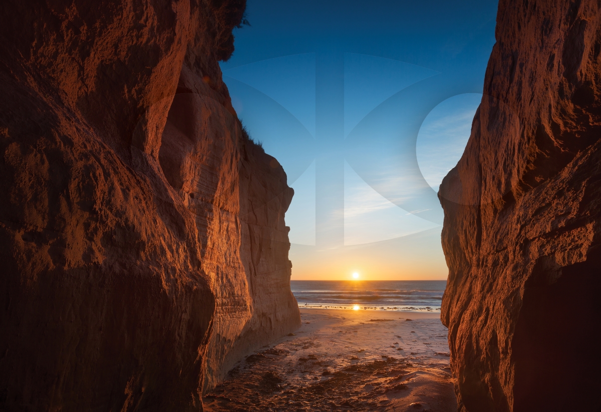 Soleil levant sur l’eau entre deux falaises rouges sur une plage des Îles de la Madeleine, au Québec, superposé d’un logo transparent du Comité consultatif autochtone.