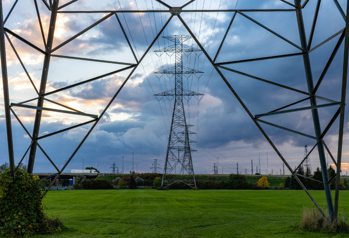 Une infrastructure de lignes de transport d'électricité à haute tension est visible en arrière-plan, encadrant un paysage surplombé de nuages sombres dans le ciel et d'autres lignes électriques au loin.