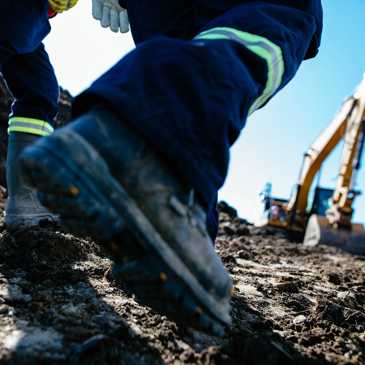 Focus on walking boots of two workers in mud. Blurred background of machinery.