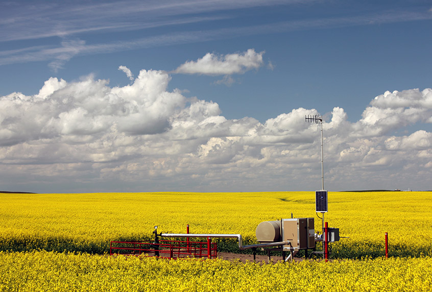 Natural gas well head in a canola field under partly cloudy skies