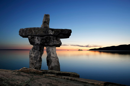 The silhouette of an inuksuk at the edge of a lake at sunset