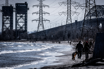 End of winter, people stroll the shore of Lake Ontario in Burlington on a sunny day, power lines and a bridge are in the distance
