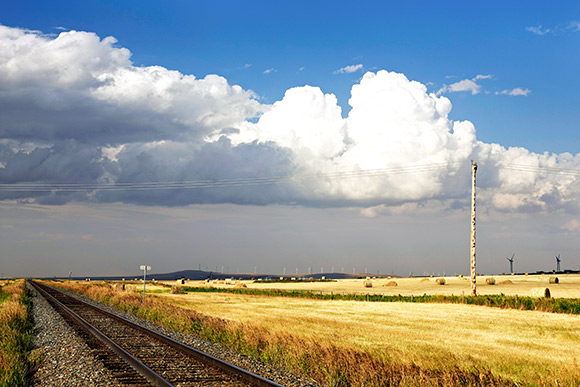 Voies ferrées défilant dans des champs de blé, dans le Sud de l’Alberta. Lignes électriques dans le ciel et éoliennes à l’horizon avec les montagnes en arrière-plan.