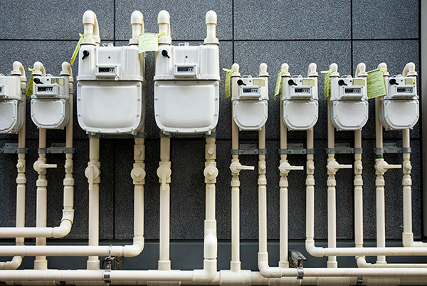 A photo of several white natural gas meters and pipelines attached to a grey brick industrial building.
