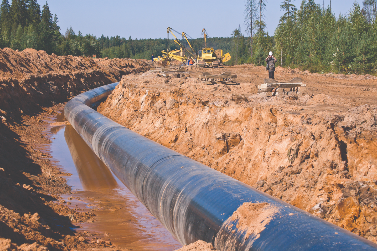 Photo of a trench dug into the ground with a length of pipeline in it. A worker in uniform is standing near the trench and construction vehicles can be seen in the distance.