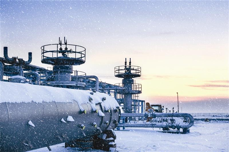 Natural gas compressor station with frost covering pipes and valves on a cold wintery day. The sun is rising in the background and there are clouds in the sky.