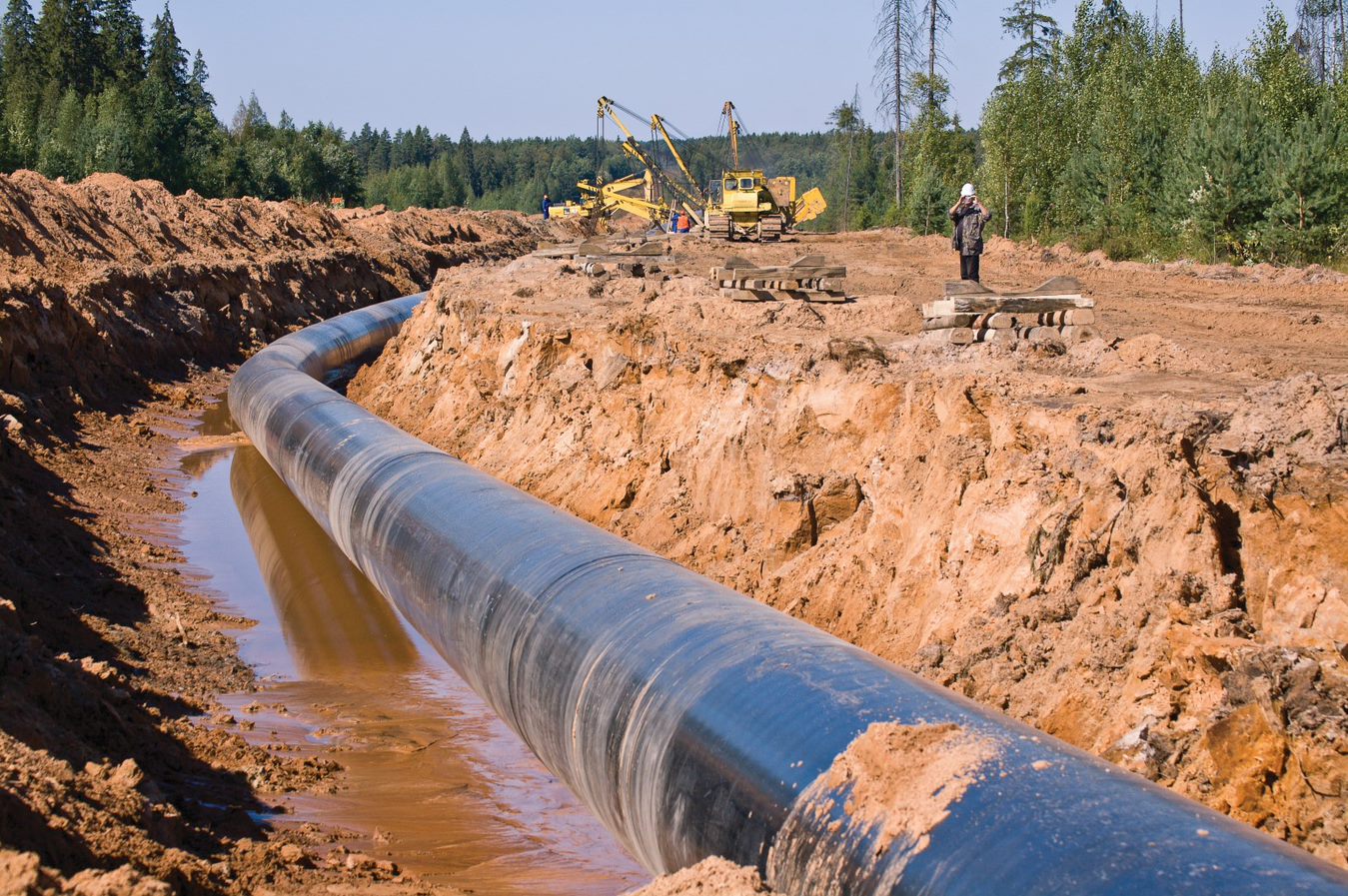 Pipeline in muddy trench with workers and heavy duty construction equipment in background.